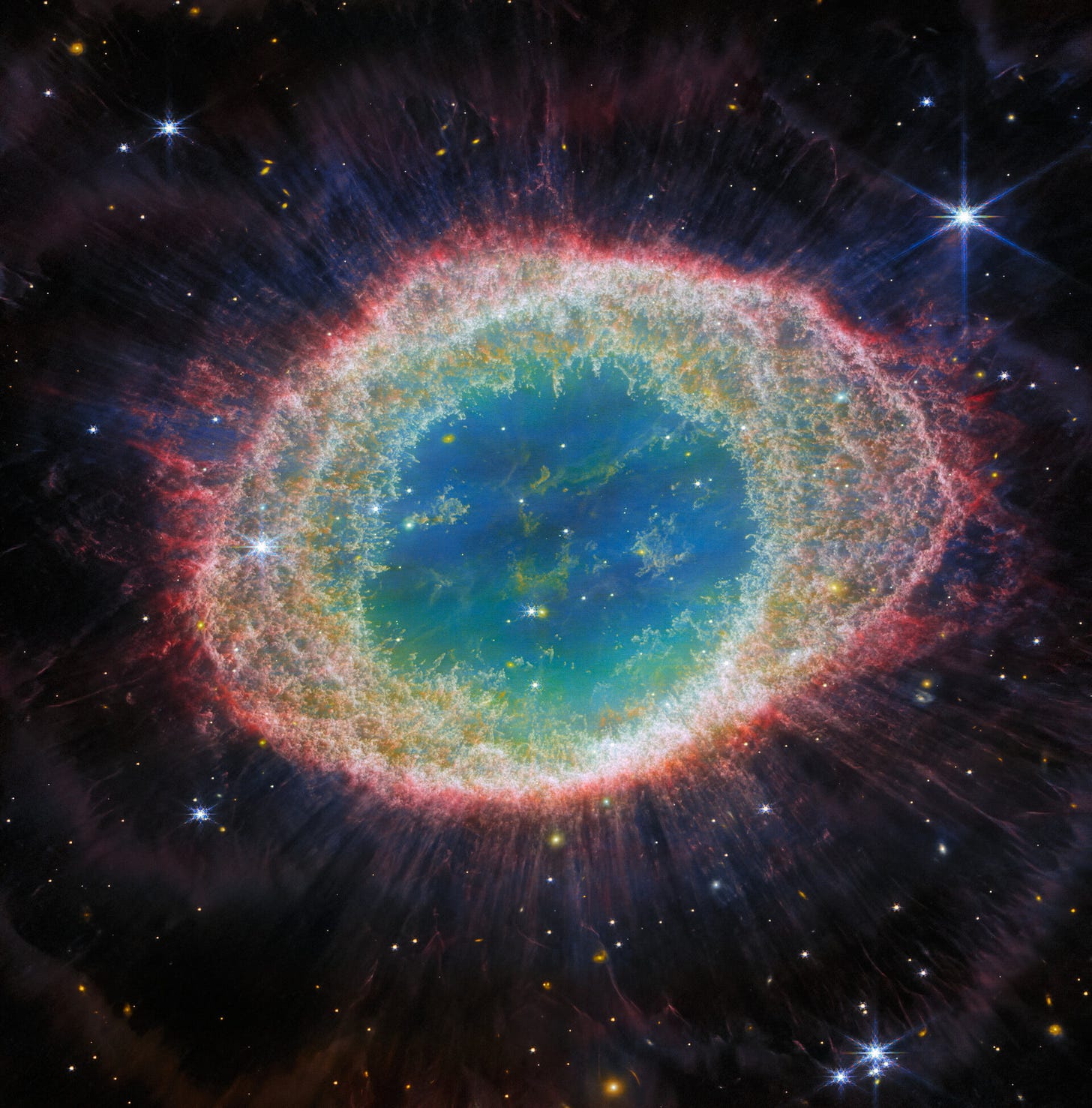 This image of the Ring Nebula appears as a distorted doughnut. The nebula’s inner cavity hosts shades of blue and green, while the detailed ring transitions through shades of orange in the inner regions and pink in the outer region. The ring’s inner region has distinct filament elements.