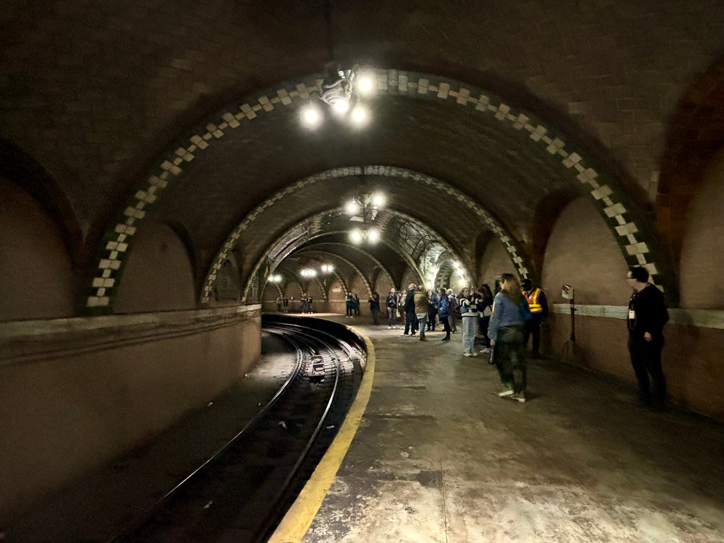 Looking down the platform of City Hall Station. There is an intricate pattern in the terra cotta tiles between the green and white tiled arches. The chandeliers are bright. There are people milling around taking pictures excitedly.
