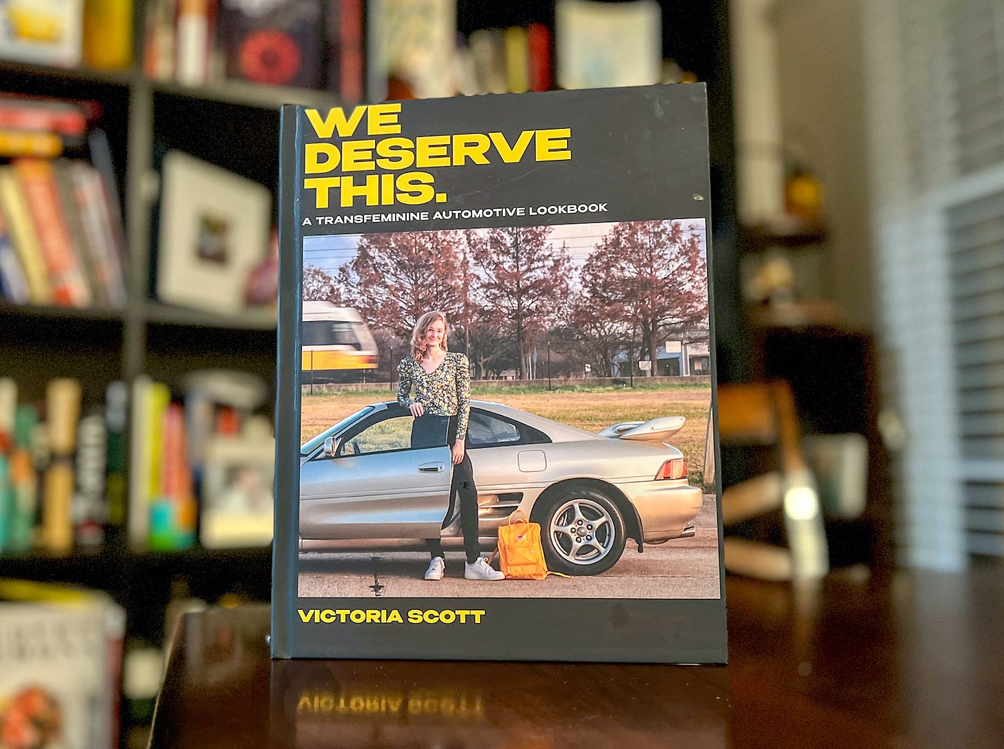 Front view of the coffee table book "We Deserve This" by Victoria Scott, standing on a table with a blurred bookcase behind it. A model poses on the cover standing in the doorway of a metallic gold Toyota MR2.