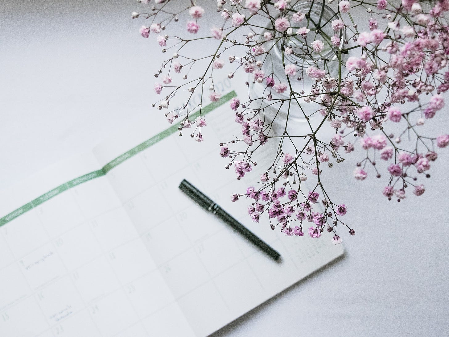 An open diary on a white table. A black pen rests on the diary. A vase of pink flowers leans over the diary.