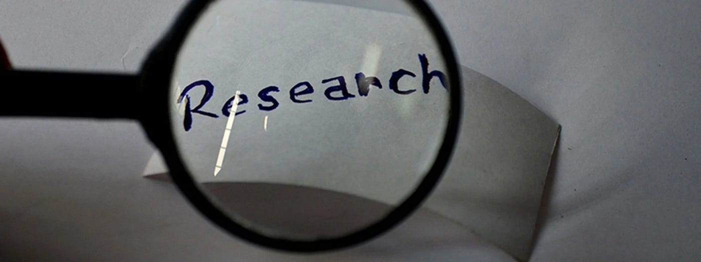 Black and white photo of a magnifying glass enlarging the handwritten word "Research" on a piece of paper.