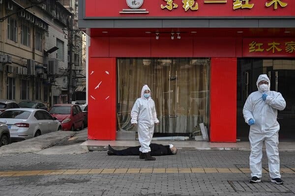 An man in black clothing lies on the cobblestone pavement outside a red building with yellow Chinese lettering. Two workers in officials in full protective gear stand nearby.