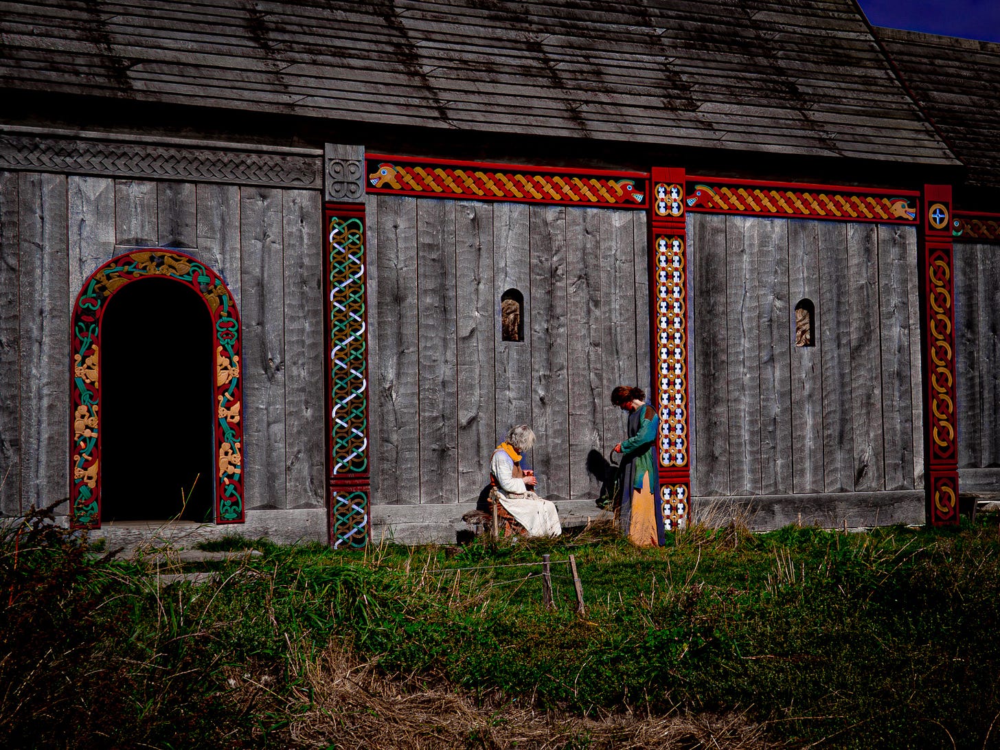 Two women in traditional Viking garb working in the sunlight outside of a replica Viking era structure with painted beams. Two women in traditional Viking garb working in the sunlight outside of a replica Viking era structure with painted beams.