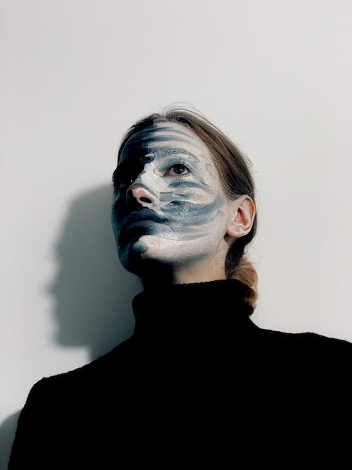 Free From below of calm strange female with paints on face wearing black turtleneck looking away while standing against white background Stock Photo