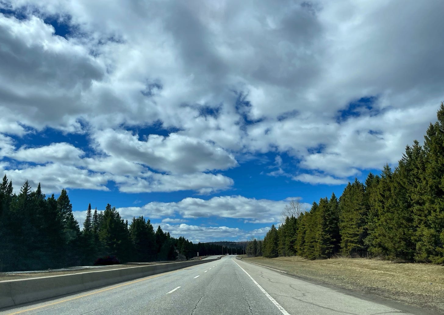 photo taken through the windshield on an empty Canadian highway with blue sky, white clouds and spur trees on both sides