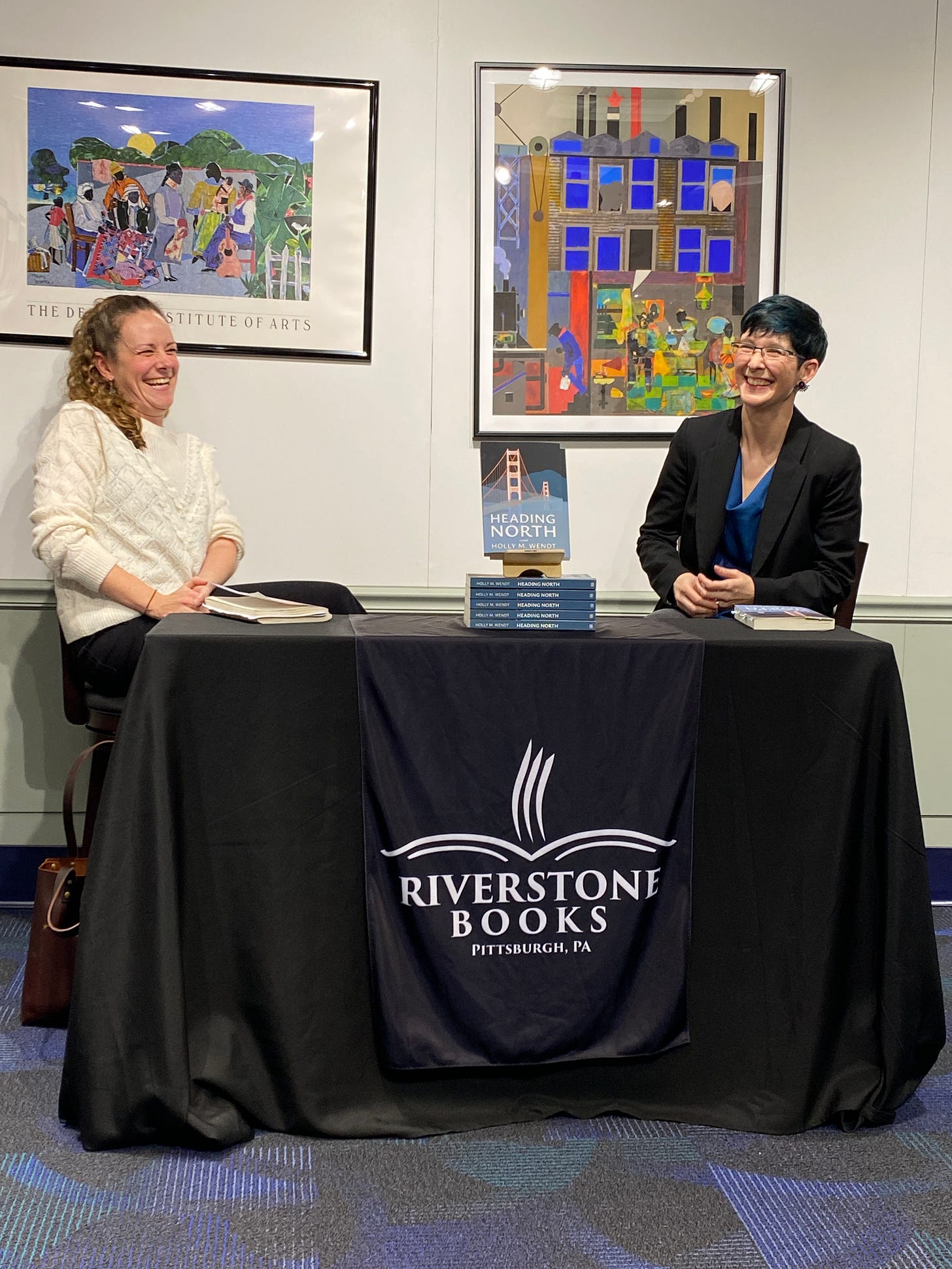 Clare Beams, left and Holly M. Wendt, right at Riverstone books, Pittsburgh, with a table between them and works of art behind them, giving an author talk and Q&A.