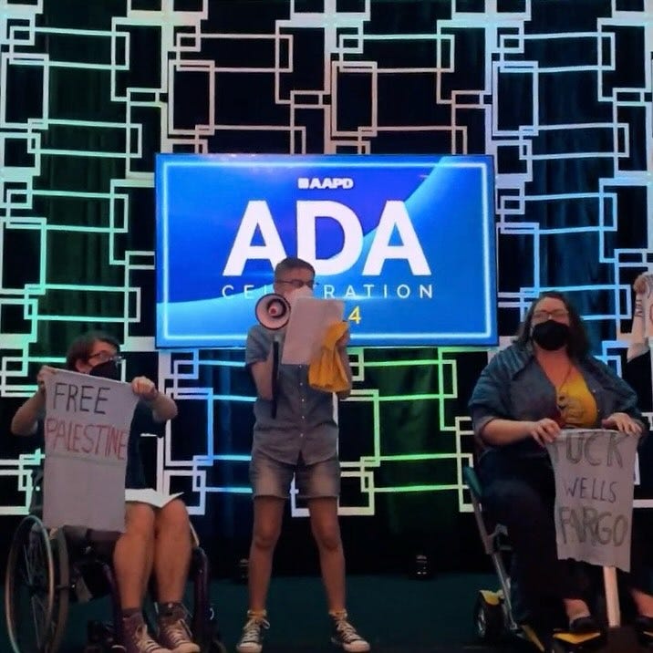 Three individuals on stage — one is in a wheelchair holding a sign that says “Free Palestine”, the second is holding a megaphone, and the third, Jen Deerinwater, is in a motorized scooter holding a sign that says, “Fuck Wells Fargo"