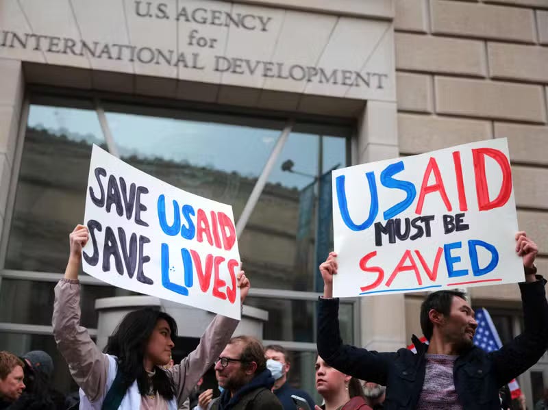 A group of protestors gather outside a building with the logo "U.S. Agency for International Development" on a building behind them. They hold signs saying "SAVE USAID SAVE LIVES" and "USAID MUST BE SAVED." A group of protestors gather outside a building with the logo "U.S. Agency for International Development" on a building behind them. They hold signs saying "SAVE USAID SAVE LIVES" and "USAID MUST BE SAVED."