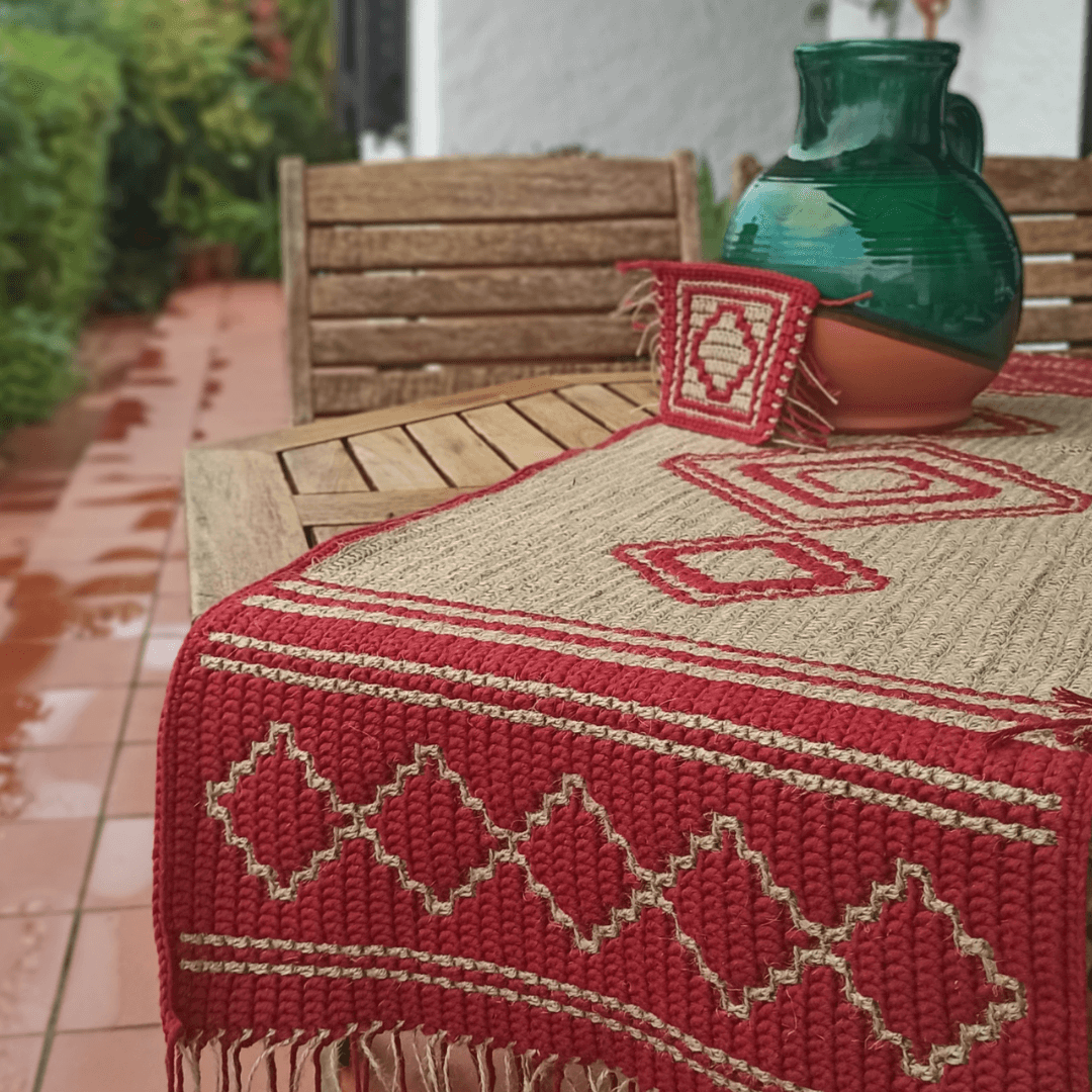 Dark red cotton and jute table runner in mosaic crochet with geometric design and fringes, on table with coaster and green glazed ceramic rustic jar.