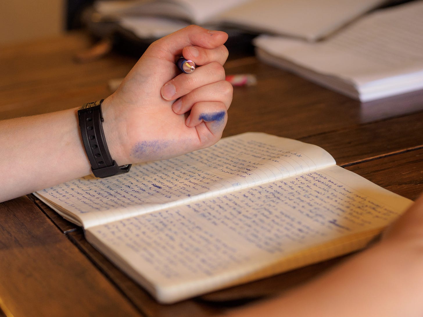 Photo of a left hand against a wooden table, writing in a notebook in blue ink. Blue ink is also smudged down the left arm.