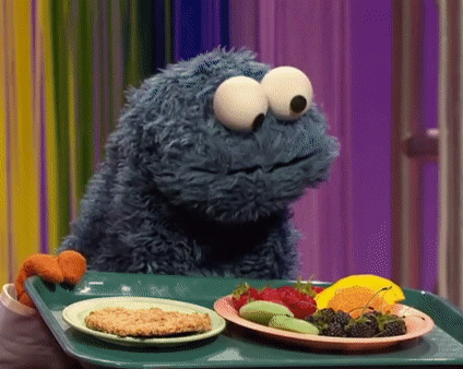 Cookie Monster trying to choose between a plate of fruit and a big cookie, looking back and forth from one to the other