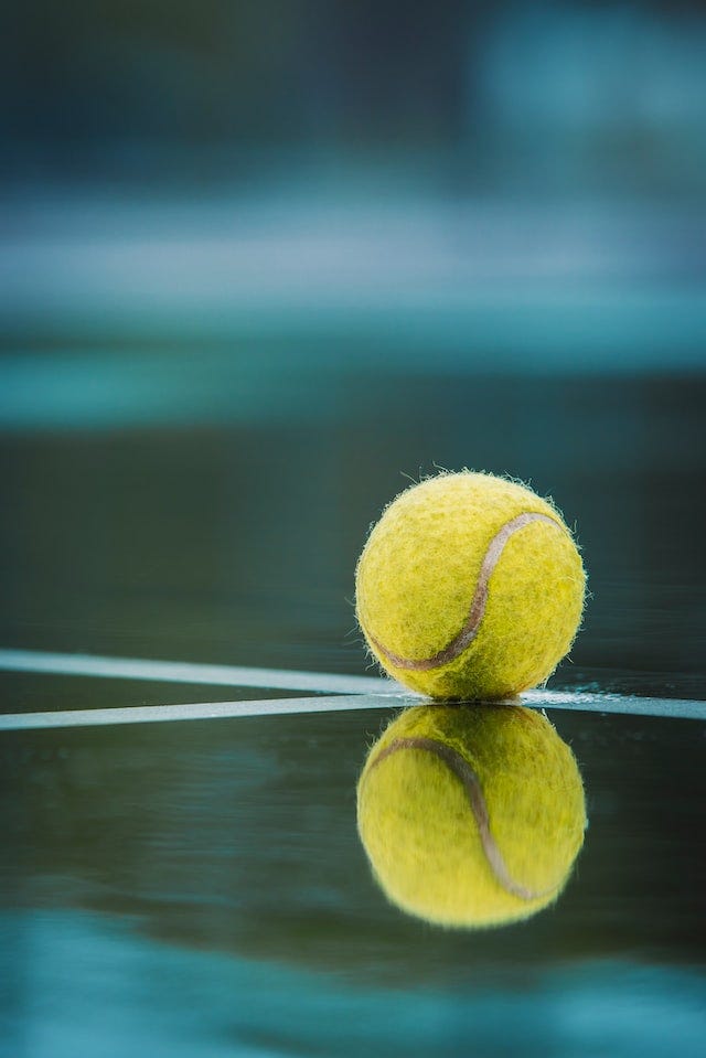 In close up, a waterlogged yellow tennis ball sits at the intersection of two court lines (the baseline and a singles line) on a wet court.