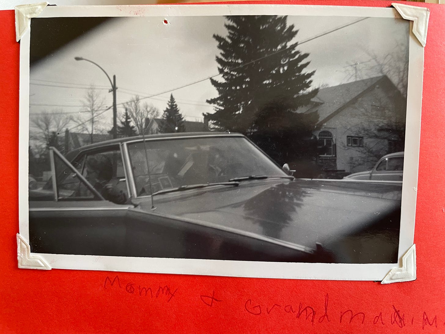 A black and white photo of a 1970s model car parked in front of a big tree. Caption, in a child's (my) handwriting says, "Mommy & Grandma M." 