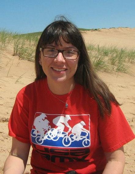 A young woman with long-brown hair and glasses, wearing a red shirt with bicycles on it, sitting on the sand dunes in Northern Michigan