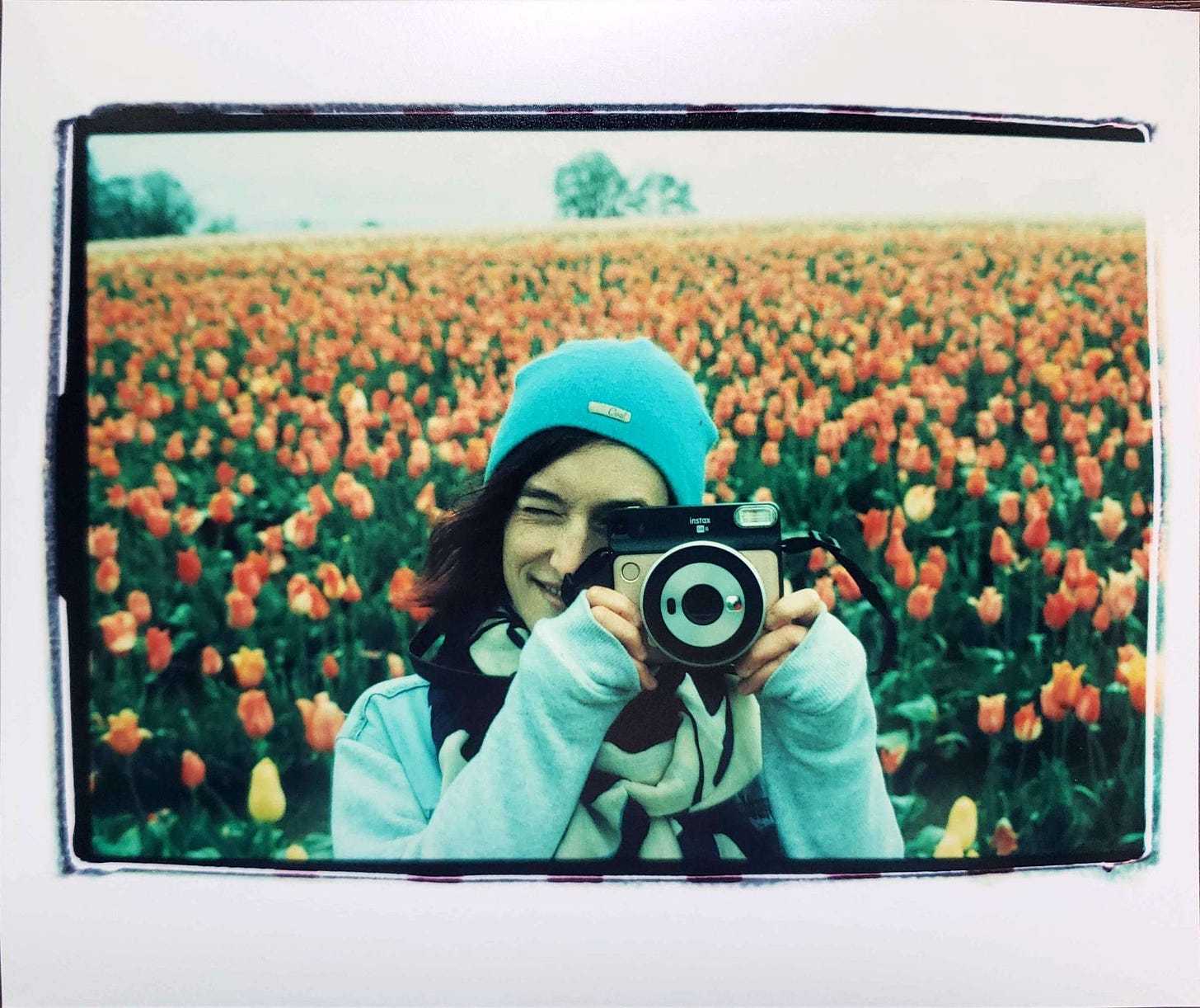 Sarah wears a blue beanie, scarf and a jacket, standing in front of a field of orange tulips They hold their camera up to their eye to take a photo, smiling.