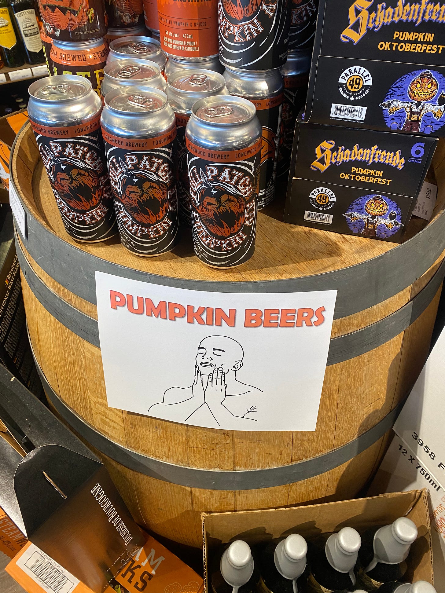 A barrel with a display of different cans and bottles of pumpkin beer. A sign taped to the front of the barrel reads 'PUMPKIN BEERS' in orange text and there's a meme beneath it, a line drawing of a bald man touching his face with his eyes shut, looking euphoric.