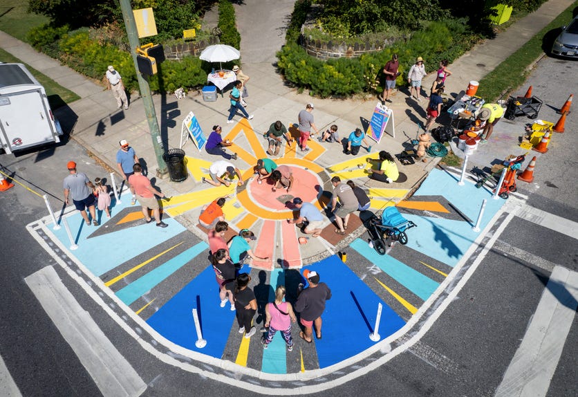 Shot from above, the photo shows a street corner being painted with a stylized, multi-colored sun design. The design covers the sidewalk corner, encompassing the width of both sidewalks that extend from the corner, and it extends into the street to the width of a parking space on both streets. About a dozen people are on the ground painting while about twenty additional folks watch.