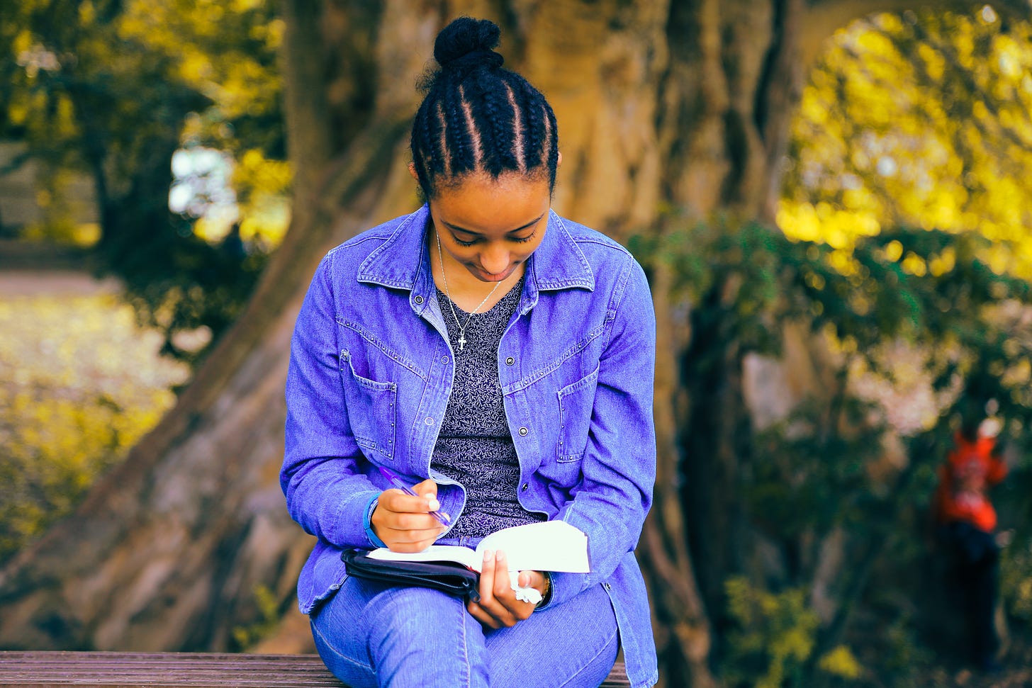 Young black woman in a denim shirt sitting on a bench outdoors, writing in a notebook.