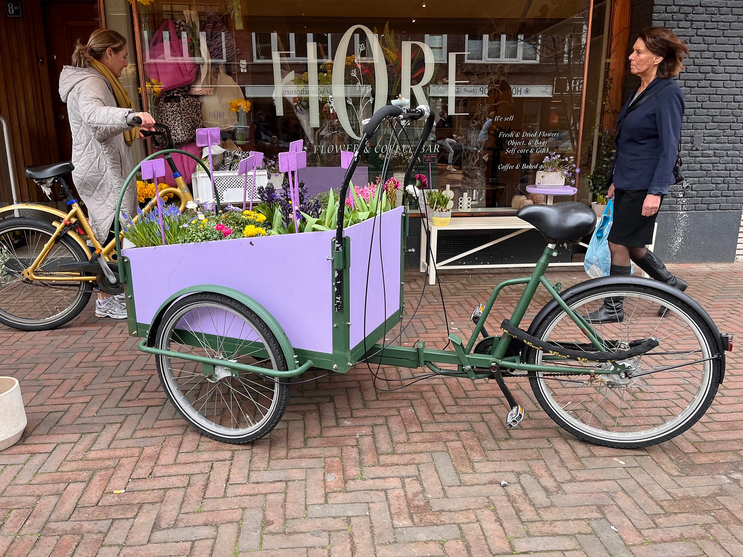 A green, three wheeled cargobike with a bright purple wooden box in the front. Inside the box are flowers for sale with price tags attached to long sticks. 