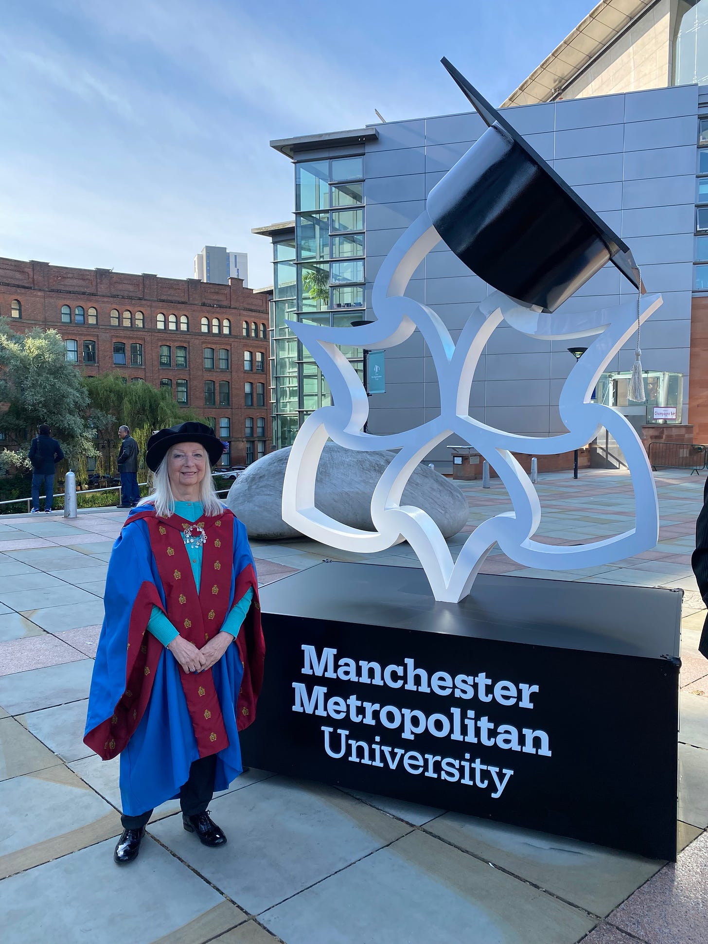 Woman wearing graduation robe and bonnet standing in front of large statue type sign for Manchester Metropolitan University with buildings in the background and a light blue sky