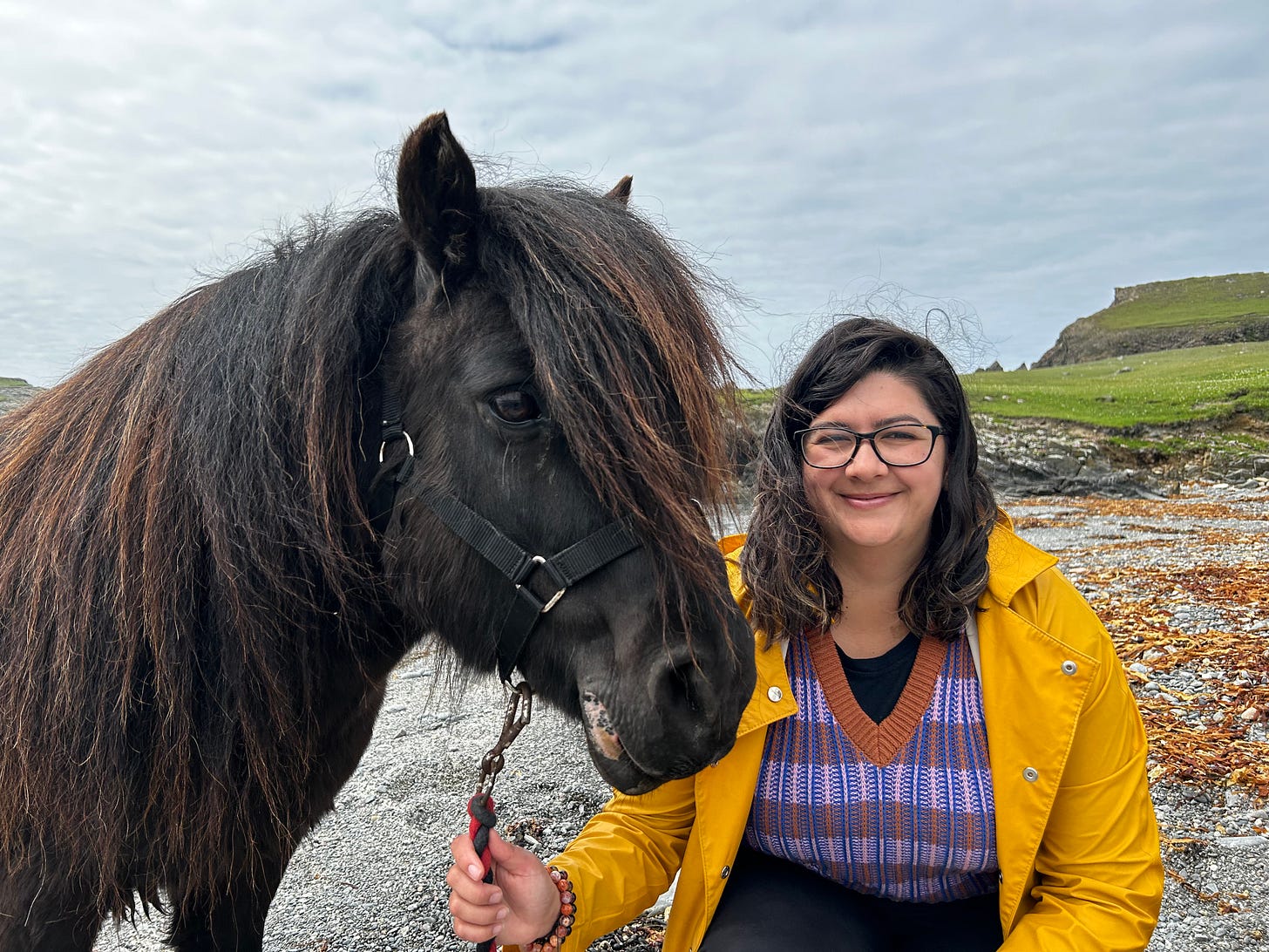 A woman in a yellow raincoat and glasses poses with a dark brown Shetland pony.