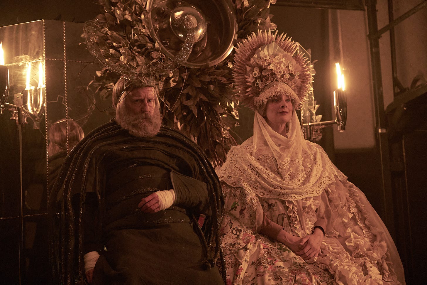 a white man with a grey beard wears a dark shroud and complicated gilded headpiece seated next to a white woman with blonde hair wearing a gown of florals and lace with a gilded headpiece resembling a sunflower. they are seated on a stage illuminated by candelabra.