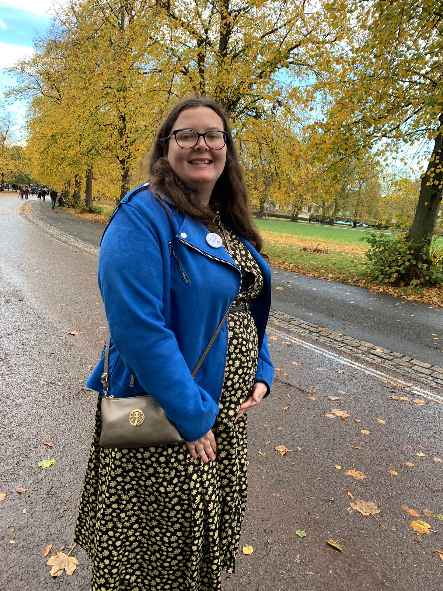 Charlotte, with brown hair and glasses, is cradling a small but growing baby bump and showing off her Baby on Board badge, with an autumnal park in the background