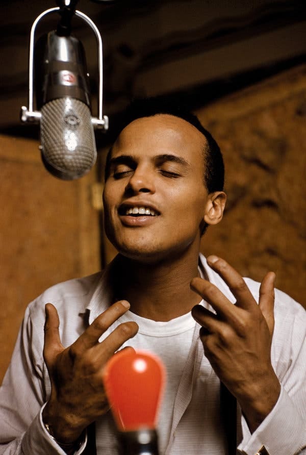 A close-up color photo of a young Mr. Belafonte singing into a suspended recording studio microphone, his eyes closed and his hands raised and gesturing. He wears a light-colored collared shirt, with cuff links, unbuttoned to his chest, over a white T-shirt.