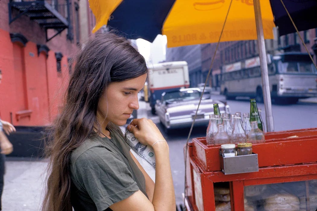 A young Bernadette with long dark hair looks down pensively as she holds a dollar bill. She is standing on a street corner in front of a hot dog cart.