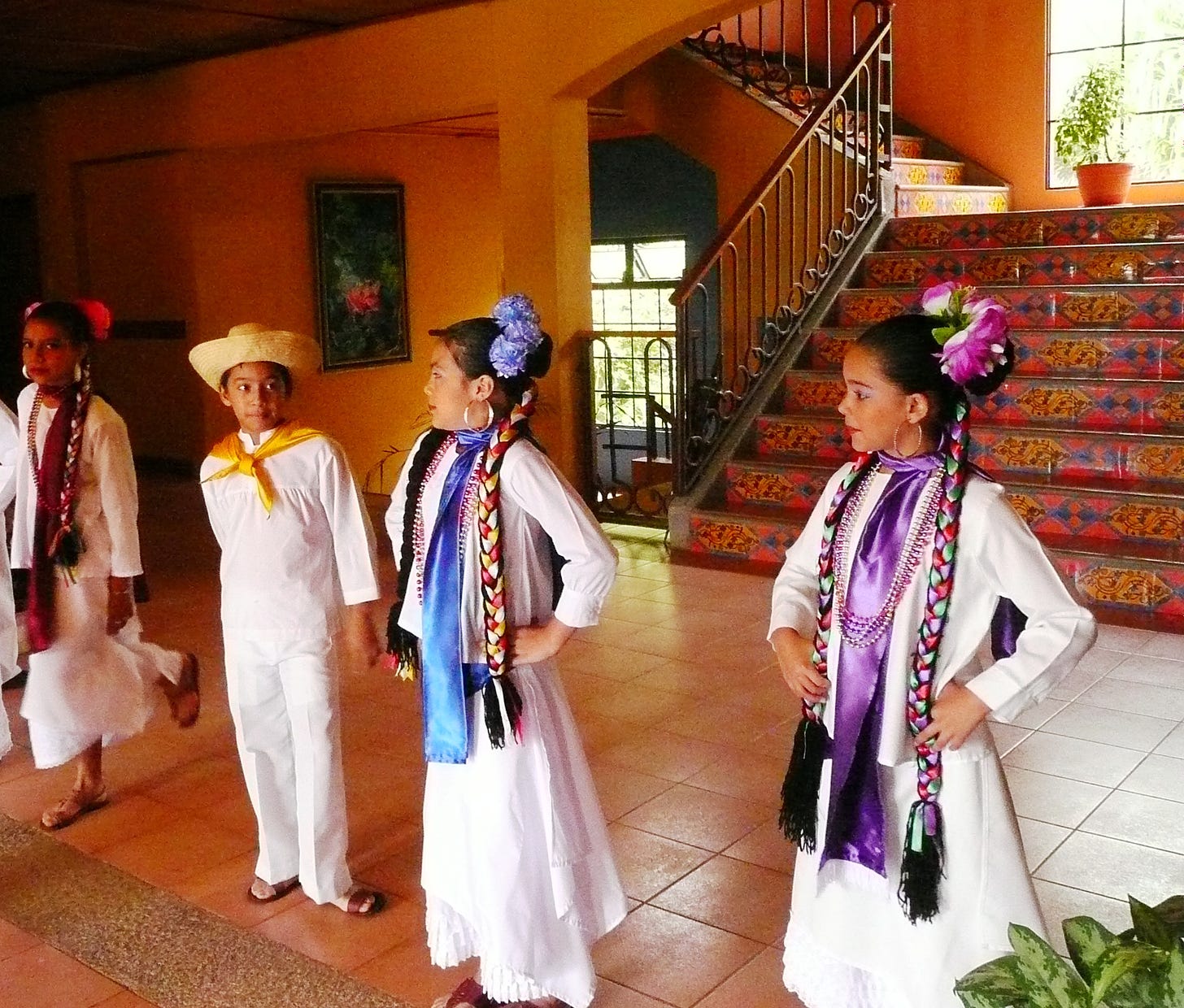 In this photo you can see young Nicaraguans preparing to perform - they are dressed in a traditional white costume and also have colourful flowers and neckties around their head and bodies. The boy wears a hat and the girls wear braids and flowers in their hair. They stand on a terracotta floor with a staircase and window behind them.