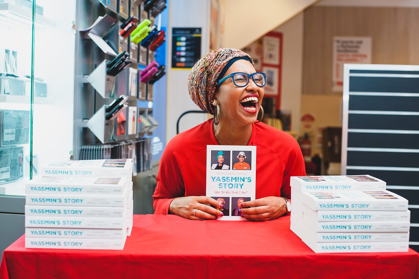 Yassmin Abdel-Magied holding a copy of her first book, Yassmin's story, sitting at a table between two piles of her books and looking off to the side, eyes closed mouth open in an extreme expression.