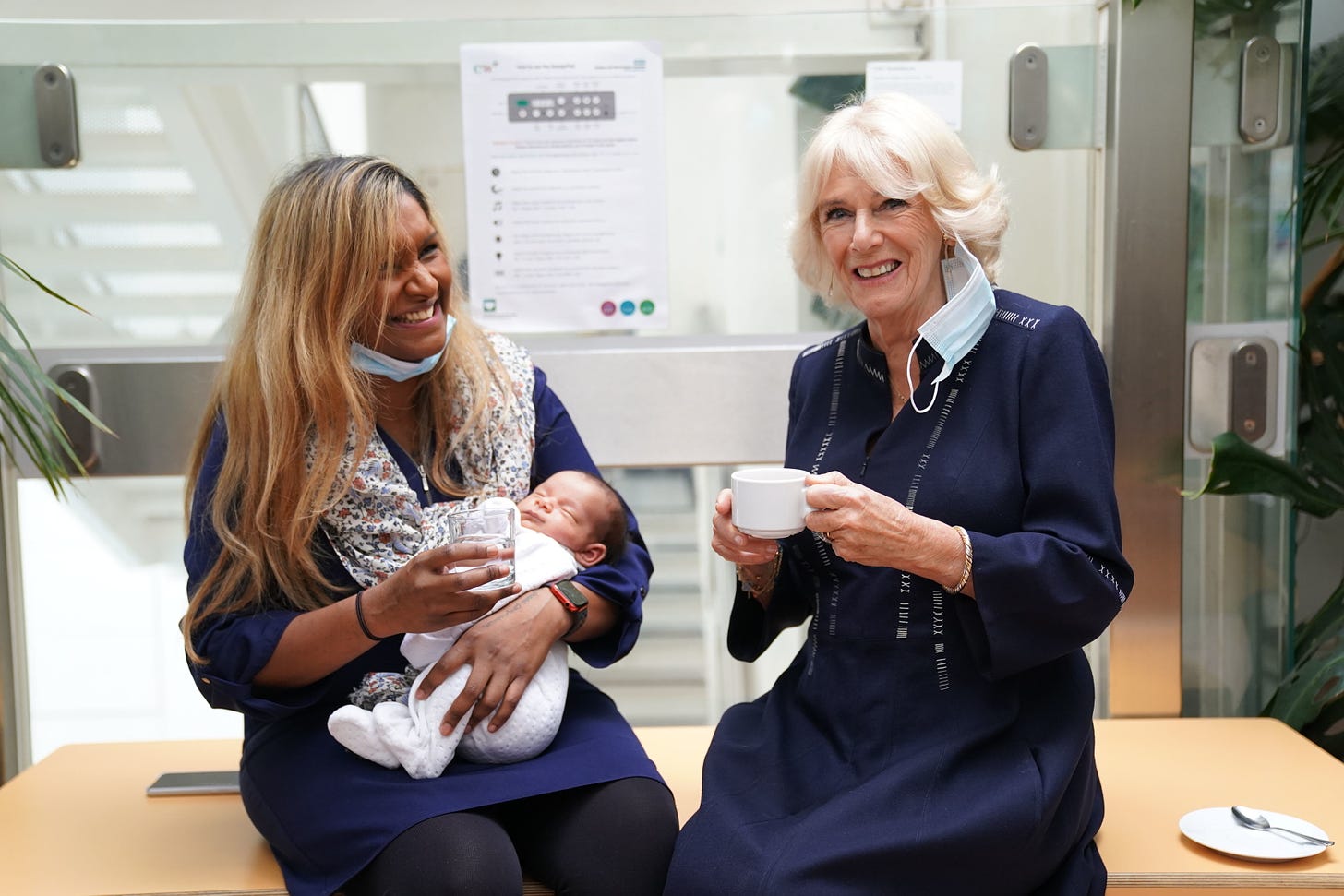 Queen Camilla smiling and holding a cup next to smiling lady holding a baby