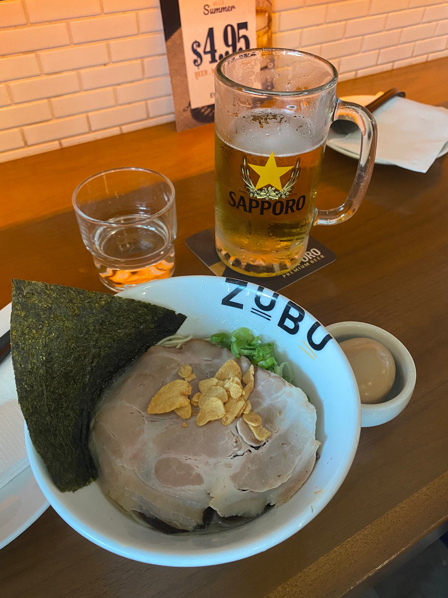 A small bowl of ramen with nori, chashu, green onion, and garlic chips. Next to it is a small dish with a seasoned egg, and a beer in a 'Sapporo' branded mug.