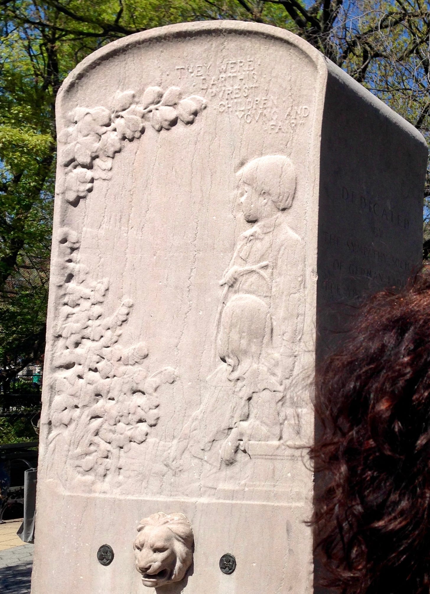 A white marble relief of a young boy and girl looking off into engraved vines. Above them is the line, "They were earth’s purest children young and fair" from Percy Bysshe Shelley's Canto VIII. A woman with a beautiful head of thick, curly hair stands directly to its right. There is a lion's head fountain mouth beneath the engraving.