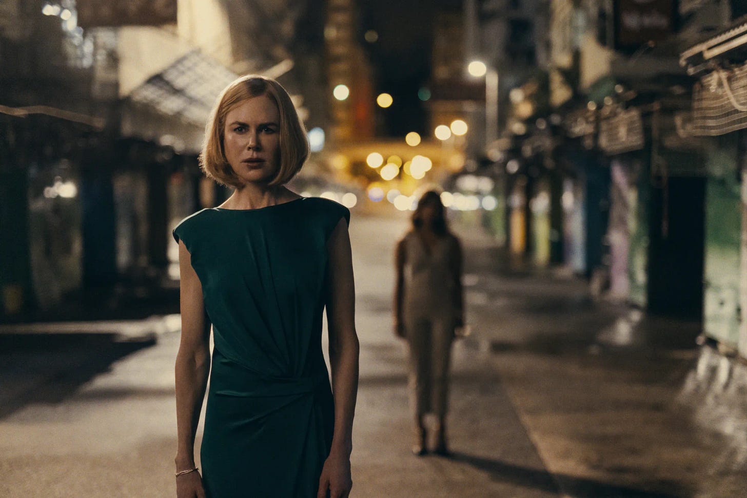 Nicole Kidman em uma rua deserta, à noite, exceto pela presença de outra mulher, em segundo plano e desfocada, atrás dela. Nicole veste um vestido verde escuro e tem cabelo na altura do queixo, loiro e liso.