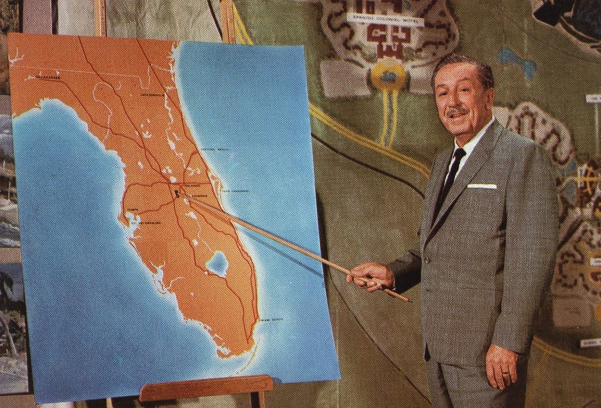 A man in a gray suit, identified as Walt Disney, stands in front of a large map of Florida, pointing at a location near Orlando with a wooden pointer. Behind him, a conceptual map of a planned development, including roadways and a central hub, is visible. The image appears to be from a historical presentation about the development of Walt Disney World.