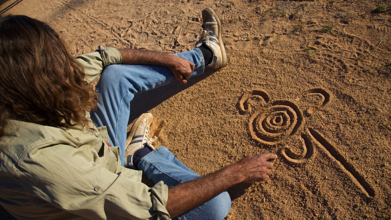 A man drawing with his finger in the dirt.