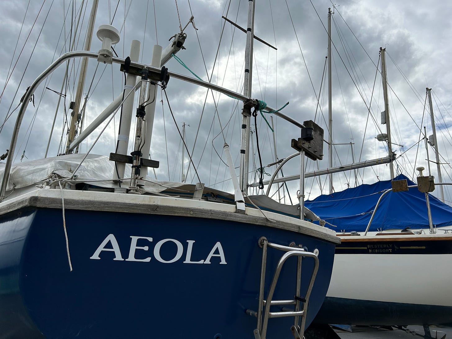 A yacht stored on dry land next to the harbour in Lyme Regis. This is the view of the stern. In white lettering on the blue-painted hull is its name Aeola. Another yacht is alongside and we can see part of its white hull. Image: Roland’s Travels.