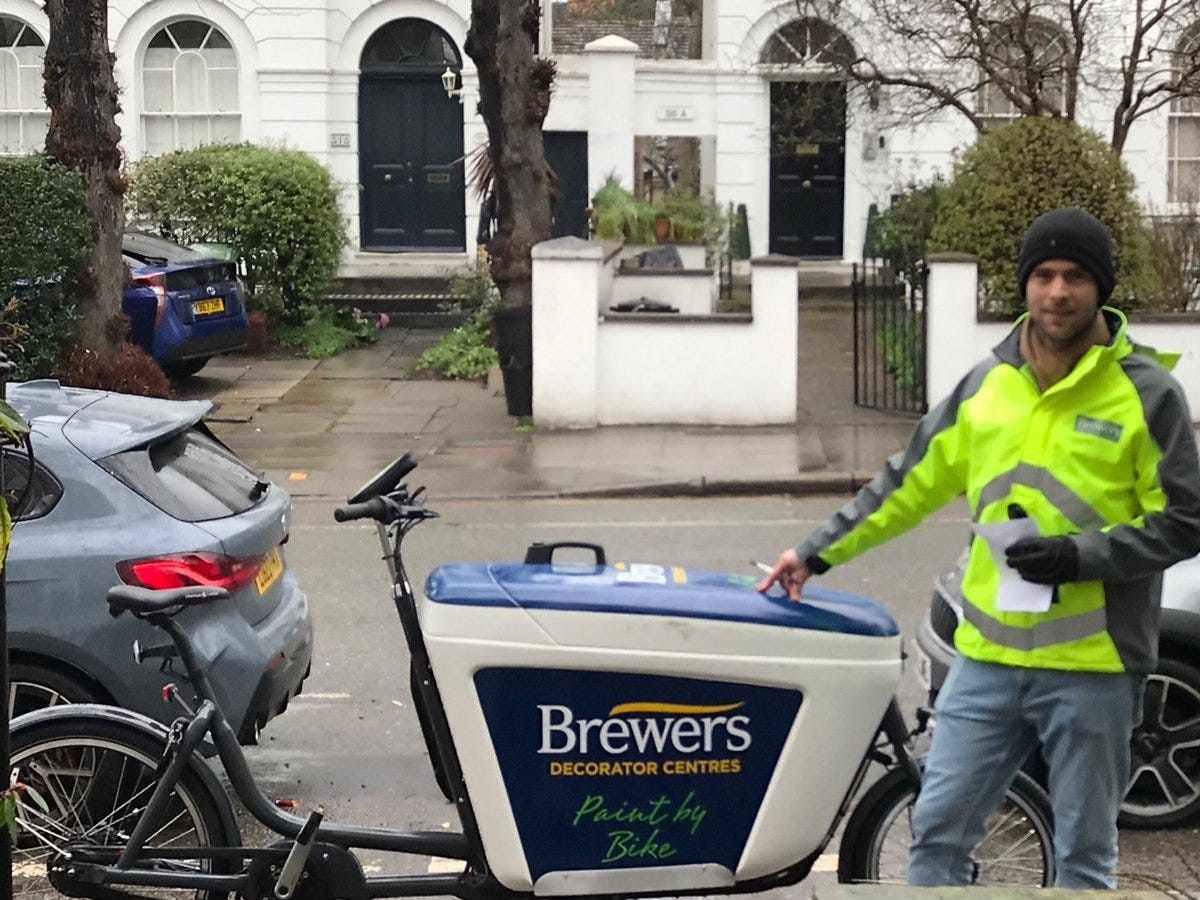 A delivery rider arrives at the destination on a residential London street. The cargobike is branded with the paint supplies company he is working for. 