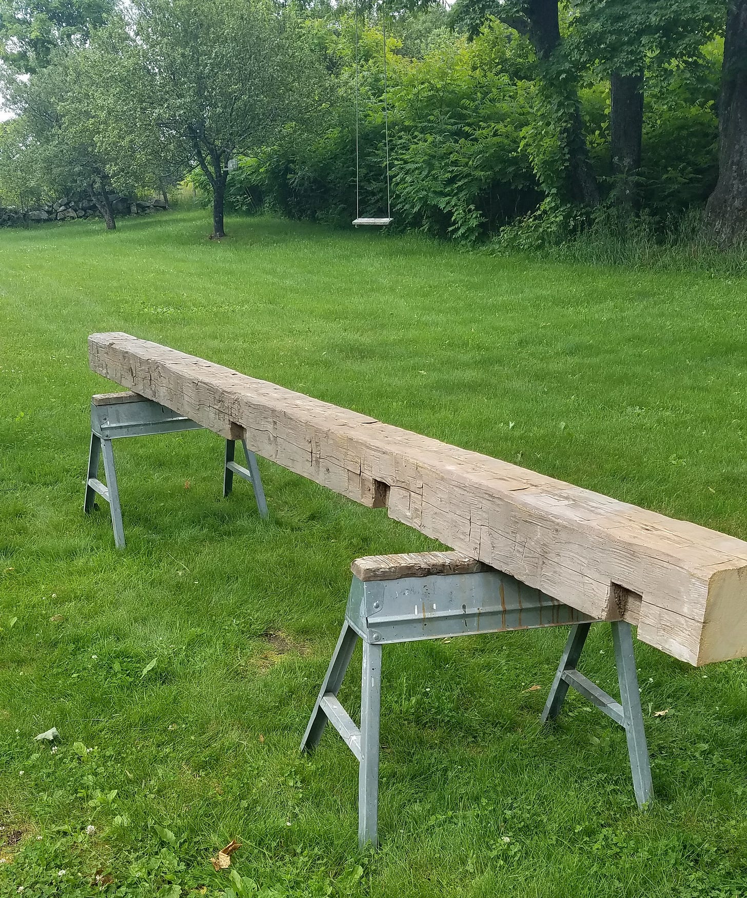 An old hand-hewn beam sits on saw horses in a yard with green grass, apple trees, and a swing