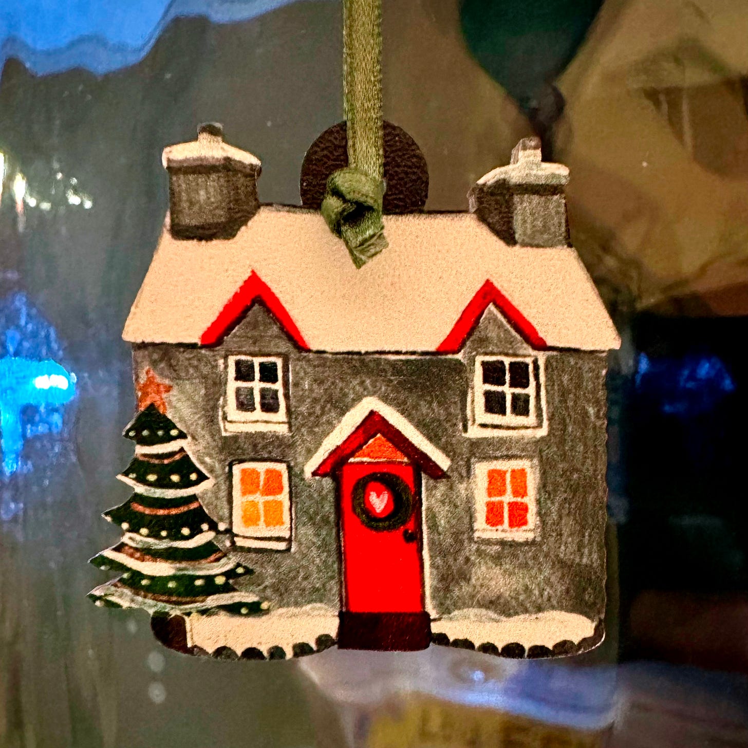 A painted wooden christmas decoration depicting a house with a snowy roof, red door and christmas tree outside.