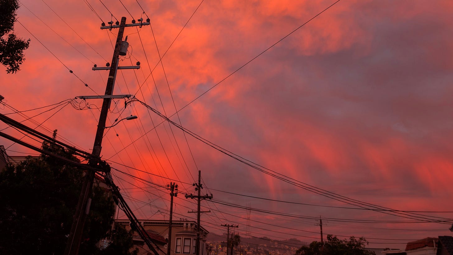 A bright pink and orange sky at sunset. In the foreground are electric poles with wires criss-crossing everywhere. In the background is Sutro Tower on Twin Peaks in San Francisco.