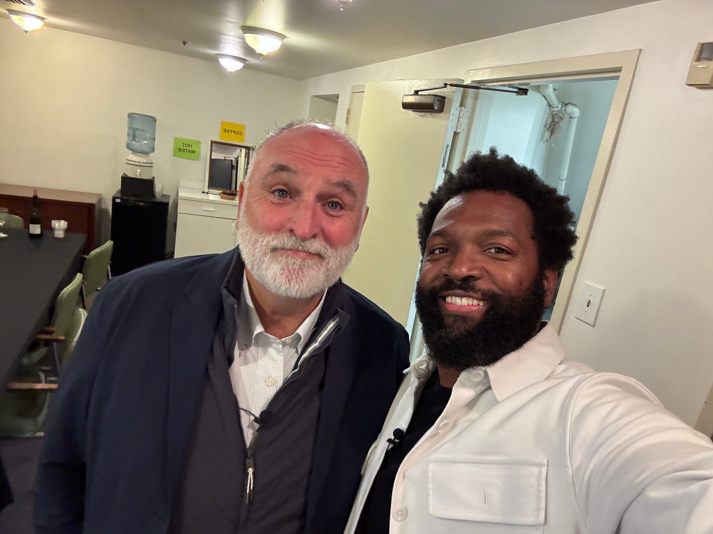 Baratunde Thurston takes a smiling selfie with Chef José Andrés in a backstage dressing room at the Japanese American Cultural Center. Behind them are folding chairs, a table, and a water cooler labeled with signs for coffee and hot water.