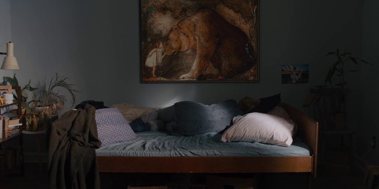 Dani lies in bed, curled up with her back to the camera. Above her is a painting of a small girl and a very large bear.
