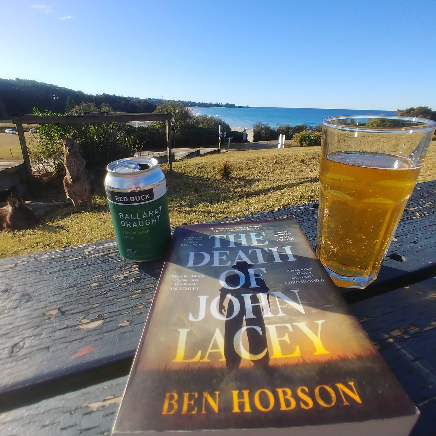 The novel 'The Death of John Lacey' by Ben Hobson, a Ballarat Draught beer from Red Duck Brewery, and a kangaroo by the beach