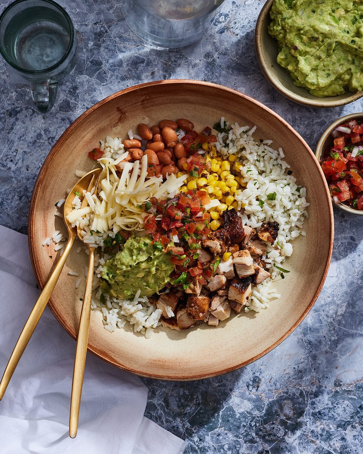 A burrito bowl sitting on top of a table filled with cheese, guacamole, diced chicken, cilantro rice, pico de gallo, and pinto beans.  