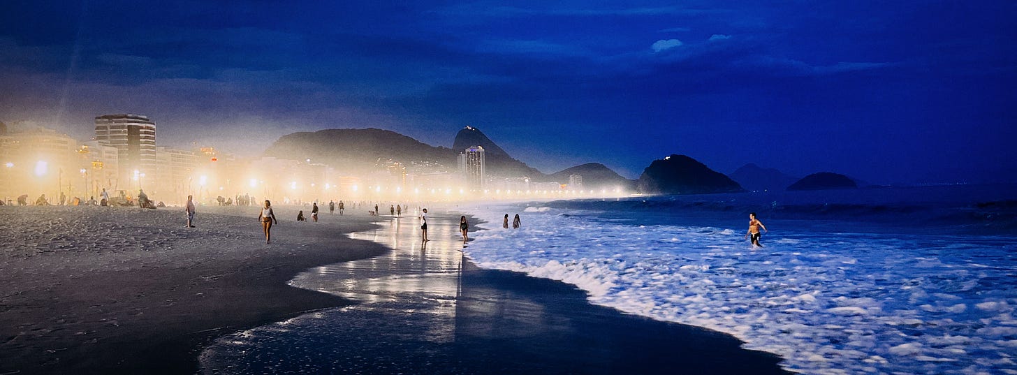 Wide angle photograph of Copacabana beach at twilight, with mountains visible in the distance and bright lights along the beach fading off into the horizon.