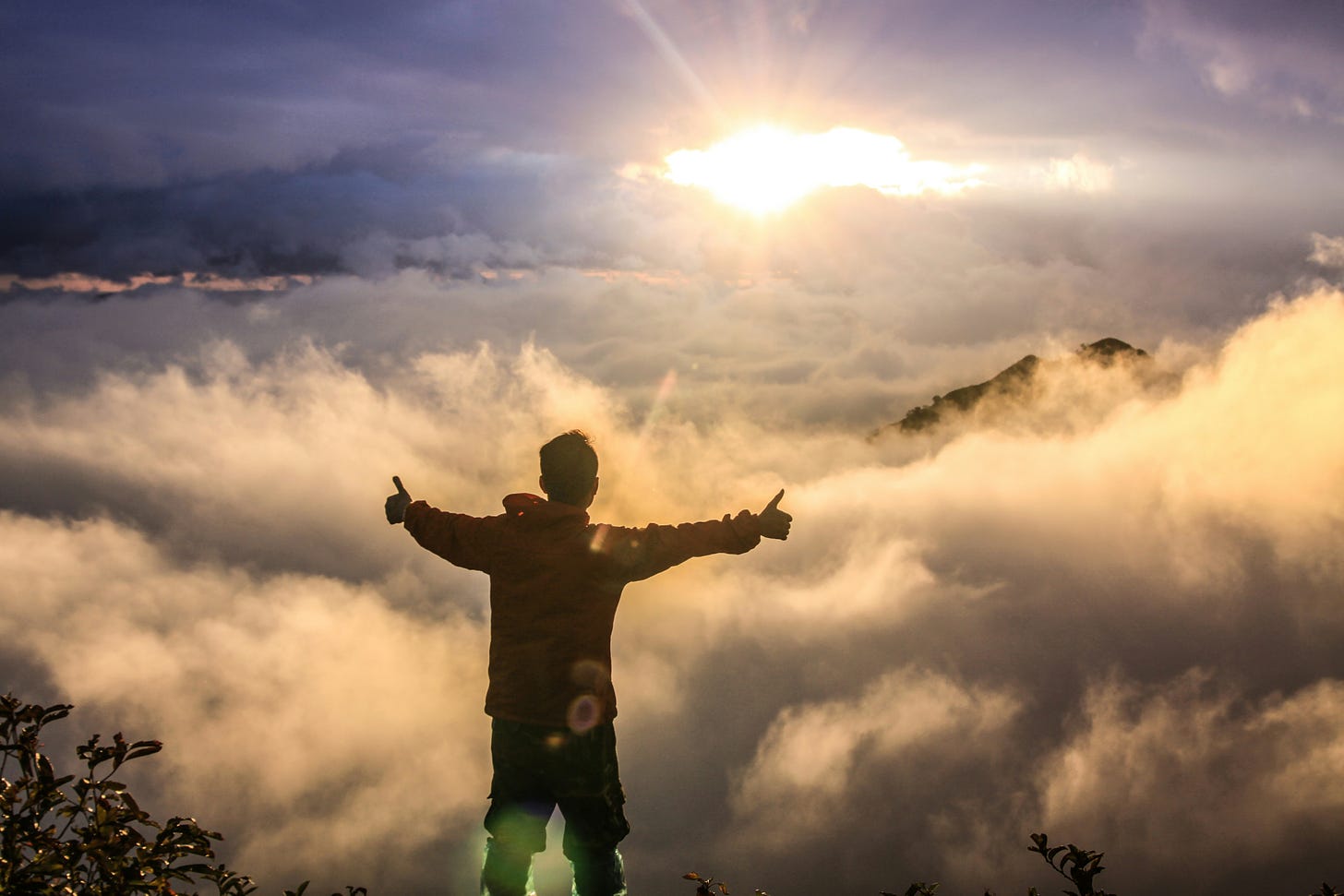 Image of a man on mountaintop with his back to the camera, arms extended and thumbs up, facing the setting or rising sun.   