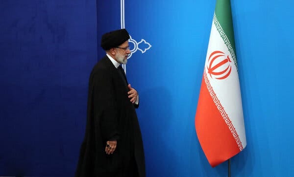 President Ebrahim Raisi of Iran wearing a black robe and a black head covering while walking on a stage next to an Iranian flag.