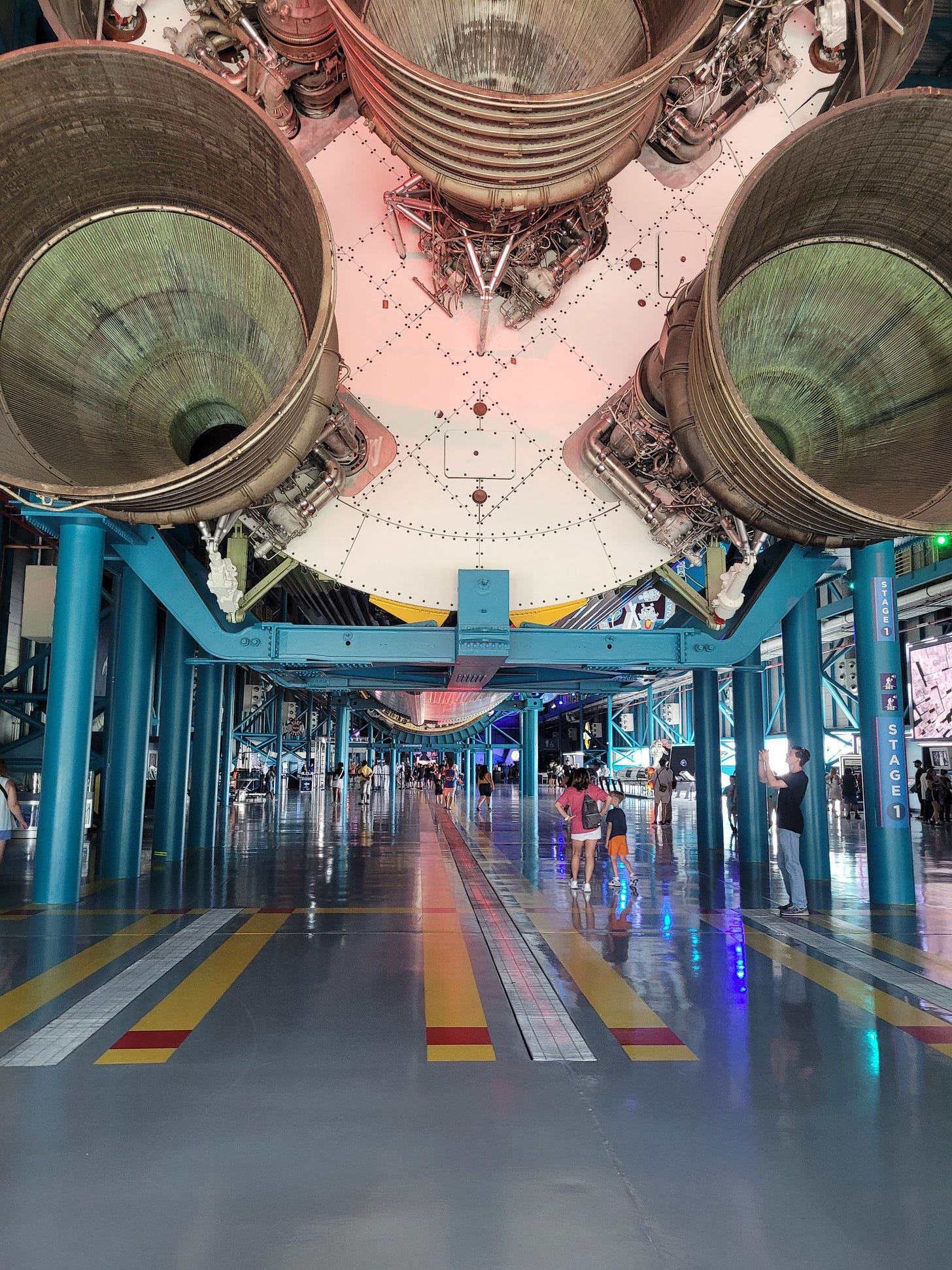 Um salão comprido, com chão cinzento com algumas faixas amarelas e uma estrutura de sustentação azul segura o foguete Saturn V deitado, numa altura em que podemos andar embaixo dele. Dá para ver as turbinas de trás e que é muito comprido.